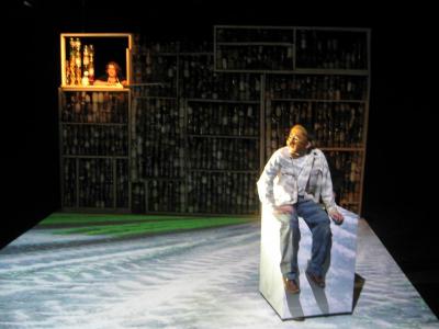 A man wearing a half mask sits on top of a washing machine. A dirt road is projected on the floor around him. Behind him a woman opens a window in a wall made of jars filled with sundry objects.