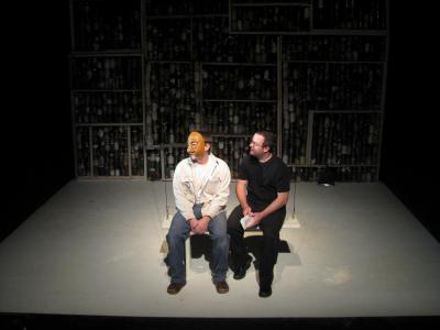 Two men, one wearing a half mask, sit on a white bench in front of a wall made of jars.