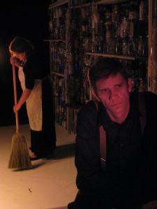 A man wearing suspenders looks troubled in the foreground. Behind him a woman wearing a white apron sweeps the floor. Behind them both is a wall made of jars filled with sundry objects.