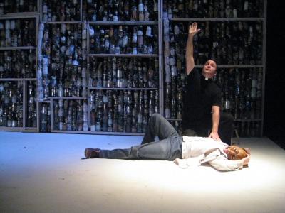 A minister kneels, reaching skyward, and places his hand on the chest of a man laying on the floor. Behind the two men is a giant wall made of jars filled with sundry objects.