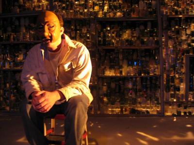 Light shines through a wall made of many glass jars. A man wearing a half mask sits in the foreground.
