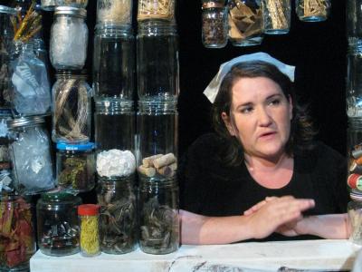 Close up of a female nurse. She’s positioned in the center of a window and surrounded by glass jars filled with sundry objects.
