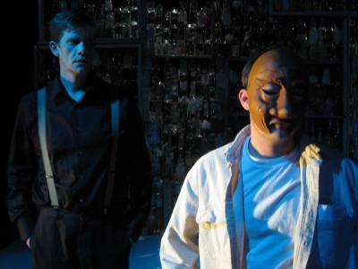 A man wearing suspenders stands behind a man wearing a half mask. Their faces are cast in shadows.