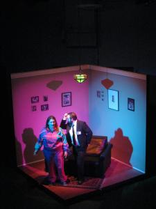 A down shot of the corner of a room, surrounded by darkness. Two people are dancing in front of a small couch. The lighting is starkly pink and blue. Photographs hang on the walls.