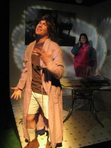A man with long hair and a bathrobe sings passionately in the foreground of a dining area. In the background, a woman in a brightly colored tracksuit looks on, confused. The lighting creates interesting shapes on the walls.