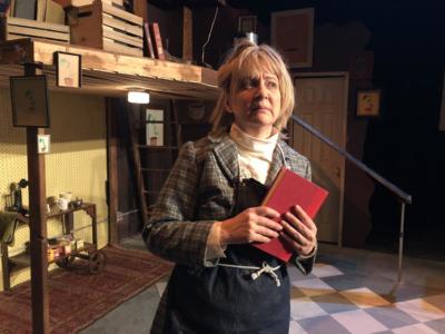 A disheveled woman wearing a while turtleneck, a plaid blazer and a blue apron is clutching a red book and looking dismayed.  She is standing in front of a door and a two level area. On the top level there are crates of books and below is clearly an area for tea and breaks.