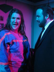 A woman in a brightly colored tracksuit sings unhappily while a bearded man in a suit watches her. The lighting is starkly blue and pink.