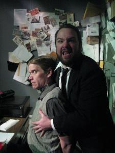 A bearded man in a suit clutches a seated man from behind, while singing angrily. The seated man looks nervous. In the background, there are a lot of letters and postcards tacked up on the wall.