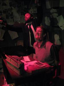 A man sitting at a desk grimaces in pain in stark pink light. Behind him, a bearded man in a suit seems to be dancing.