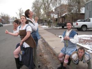 Five people in ridiculous, homemade Shakespearean outfits are by the side of a road. One sits on another. One is trying to hitch a ride.