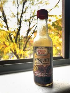 A bottle of Mesquite Liquid Smoke sits on a window ledge. Through the window behind it is an autumn tree with yellow leaves.Not only the oldest item in our fridge, but smells of burnt wood and just a hint of mystery.