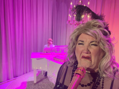 In the foreground, a sobbing blond woman is at a gold microphone. In the background, a man in a white suit sits at a white baby grand piano. The stage is washed with bright pink light.