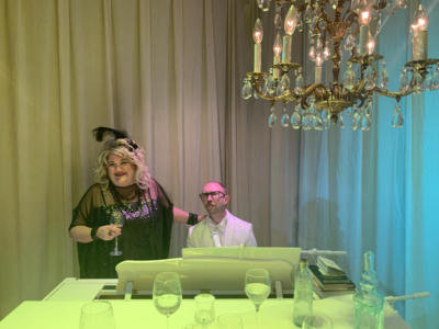 A blond woman in a black sparkly dress smiles, holding a glass of wine. A mustachioed man in a white suit and glasses sits next to her, at a piano. In the foreground is half of the piano, littered with wine glasses and book. Above is a crystal chandelier. The curtain behind them is lit in greens and blues.
