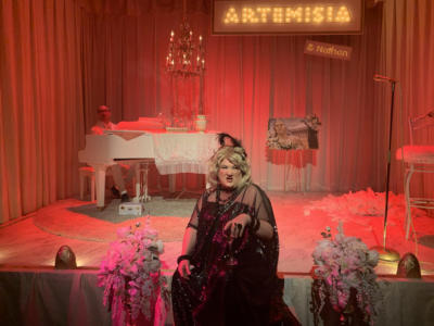 A blond woman in a black shiny dress and sunglasses sits on the edge of the stage, flowers on either side of her. She is scowling and holding a glass of wine. In the background, a man's head peeks above a white baby grand piano with a chandelier over it. The stage is lit in bright reds and pinks.