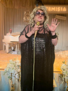 A blond woman in a black shiny dress and sunglasses stands smiling with a gold microphone. In the background, a man's head peeks above a white baby grand piano with a chandelier over it. The stage picture, with curtains and flowers, is all white.