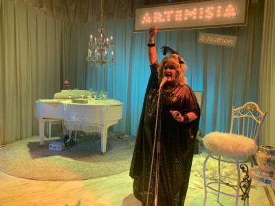 A blond woman in a black shiny dress stands at a gold microphone, holding her right arm up in the air. In the background, a man's head peeks above a white baby grand piano with a chandelier over it. Along the back is a large lit up sign that says “Artemisia” with a tiny lit up sign underneath that says “and Nathan”. The stage is lit in yellows, blues, and greens. The woman is lit in orange.