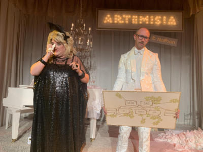 A blond woman in a black shiny dress dabs her eye with kleenex. Next to her, a man in a white suit stands holding a sign with a family tree on it. He looks annoyed. Along the back is a large lit up sign that says “Artemisia” with a tiny lit up sign underneath that says “and Nathan”.