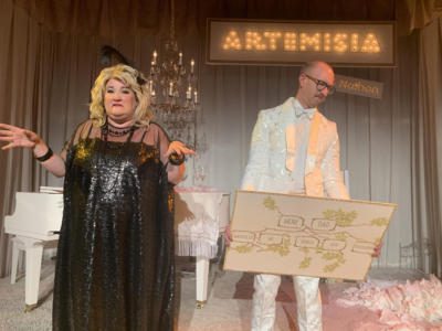 A blond woman in a black shiny dress dabs is gesturing and making a sour face. Next to her, a man in a white suit stands holding a sign with a family tree on it. He looks annoyed. Along the back is a large lit up sign that says “Artemisia” with a tiny lit up sign underneath that says “and Nathan”.