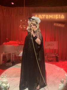 A blond woman in a black shiny dress and sunglasses sings into a gold microphone, holding a feather fan behind her head. In the background, a man's head peeks above a white baby grand piano with a chandelier over it. Along the back is a large lit up sign that says “Artemisia” with a tiny lit up sign underneath that says “and Nathan”. The background is flooded with red light