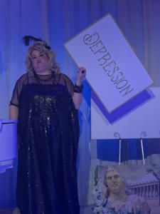 A blond woman in a sparkly black dress stands looking unhappy. She is holding a white sign that says “depression” in sparkly gold letter. The lighting is blue.