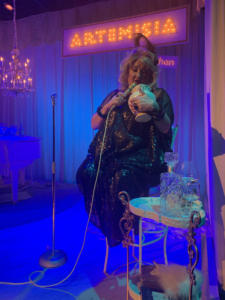A blond woman in a black shiny dress sits on a chair, singing into a microphone and gazing into an urn. In the background is a chandelier. Along the back is a large lit up sign that says “Artemisia”. The stage is lit in yellows, blues, and greens. The lighting is moody, with deep blues and violets.