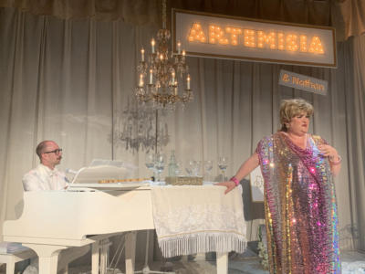 A woman with short blond hair and a very sparkly dress stands at white baby grand piano that is covered in wine glasses. A man with a mustache and glasses sits at the piano. A white curtain surrounds them. A chandelier hangs above. Along the back is a large lit up sign that says “Artemisia” with a tiny lit up sign underneath that says “and Nathan”.