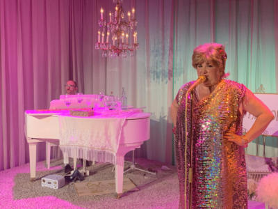 A woman with short blond hair and a very sparkly dress stands at a gold microphone. In the background, a man with a mustache and glasses sits at a white baby grand piano. A white curtain surrounds them. The lighting is a mix of bright pink and light turquoise.