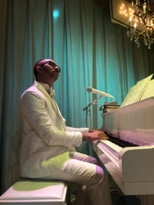 A man with a mustache and glasses sits at a white baby grand piano, his hands on the keys. He wears a white suit. The lighting is moody, with a turquoise light behind the back white curtain and some pink light shining down on the man.