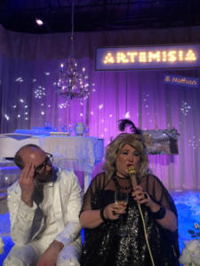 A blond woman in a black shiny dress talks into a gold microphone, while holding a glass of wine. Next to her, a man in a white suit is looking at her, exasperated. In the background is a white baby grand piano with a chandelier over it. Along the back is a large lit up sign that says “Artemisia” with a tiny lit up sign underneath that says “and Nathan”. The stage is lit with deep blue and lavender sparkly light.