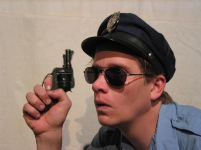 Close up on a dopey looking cop wearing sunglasses and holding his six shooter up to his face.