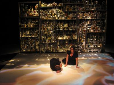 Light shines through a wall made of many glass jars. A couple lays in front of it. Flames are projected on the floor around them.