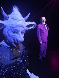 Lit in blue light, a woman dressed as a cow in a dress looks coyly at the camera. She wears a cow bell around her neck. In the distance, an older man in a magenta suit looks on.