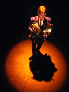 Looking down on a man in an orange spotlight. The man has many pairs of ice skates draped over his shoulders.