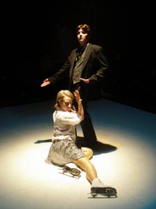 A man wearing a dark suit poses in a spotlight. Draped on his leg and across the white floor is a woman in a white shirt, sequined skirt, and ice skates.