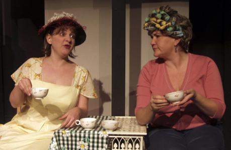 Two women holding teacups gossip at a small cafe table. One wears a hat, the other is in curlers.