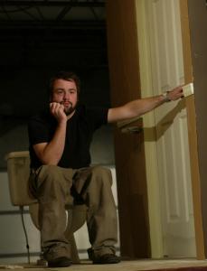 A man with a beard sits on a toilet. He is holding the bathroom door shut and has a defeated expression on his face.