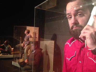 Close-up on an unhappy bearded man holding a telephone. He is behind plexiglass. In the distance are three other people sitting in large plexiglass boxes.