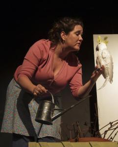 A woman in a pink top and flowered apron holds a watering can in one hand and a white cockatiel bird in the other. She is speaking to the bird, dotingly.