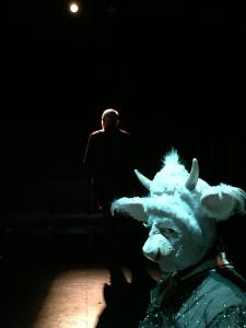 In very stark lighting a woman dressed as a cow side-eyes the camera. In the background a man is standing, but is lit from the other side so only an outline of his head and shoulders is visible.