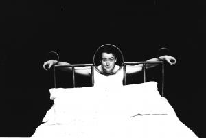 A man is pictured standing behind the headboard of a large bed. The man has positioned himself with hands and head pushed through openings in the headboard to make it seem like he’s been locked in a stockade.