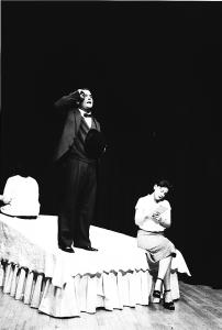 A man wearing a full suit stands on one corner of a large bed giving dictation to a woman seated on the other corner of the bed. A third woman sits at the top of the bed with her back to the camera.