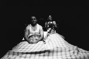 A man and woman are in bed. The man is sitting up. The woman is laying back and seems to be daydreaming. Behind her another woman, in her underwear, is wearing a fancy hat and drinking tea.