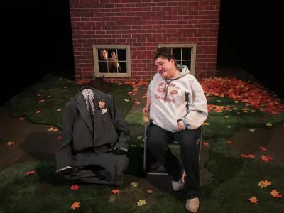 A woman wearing a sweatshirt is sitting on a folding chair in her yard.  Next to her is a men’s suit that looks like it is moving on its own.  There is no body in the suit, but it is sitting cross-legged next to her on the ground.  In the background, in the basement window of the house we can see a man looking horrified.