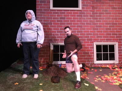 A frustrated woman stands there in a sweatshirt cinched around her face.  Her brother is kneeling down on the other side of a hole that has been dug into the ground.  He is only wearing his boxers and a tight brown turtleneck. He is clearly trying to explain himself.
