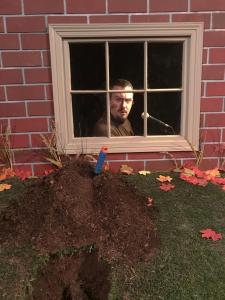 A close up picture of a hole in the ground that was dug up in the yard with a spade stuck in it.  Behind the hole is window to the basement.  In the window, we see the brother with pod cast stuff and goatee and he looks surprised.