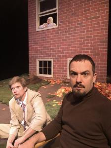 In the foreground, a man with a goatee and a tight brown turtleneck is looking intensely into the camera.  Seated next to him on the ground, his best friend is sitting there in a tan suit looking out away from the house.  Behind them both, in a window above them, the sister is looking out the window with sadness.