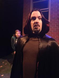 A man with an icky goatee is in the foreground looking out in a spooky way.  He is wearing a shiny black hooded cape that is red on the inside.  Behind him, his best friend is crouched down and looking as if egging his friend on.