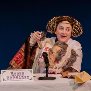 A woman dressed in 15th Century clothing and holding a mans severed head is sitting at table drinking a glass of wine. She looks as if she is on a panel of sorts. In front of her is a microphone and name tag that says “Queen Margaret”.