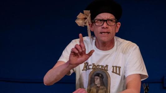 A man wearing a black beret with a giant gold bow and a t-shirt that has a picture of Richard the Third on it is holding up his index finger as if he was trying to make a point.