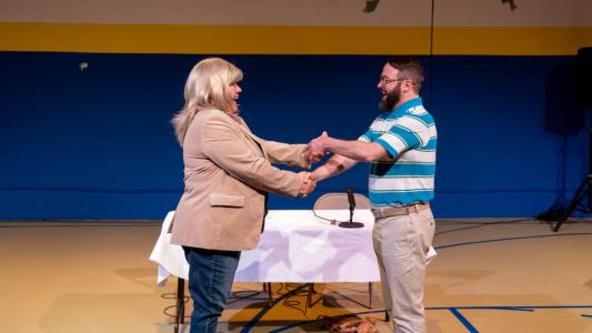 A blond lady and a man stand facing each other in front of a table. They are shaking eachothers hands with both hands crossed over the other one as if they are about to do a silly and complicated handshake.